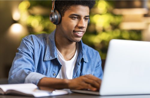 Smiling african american teen guy in headphones looking at laptop, studying foreign language through video conference application, cafe interior